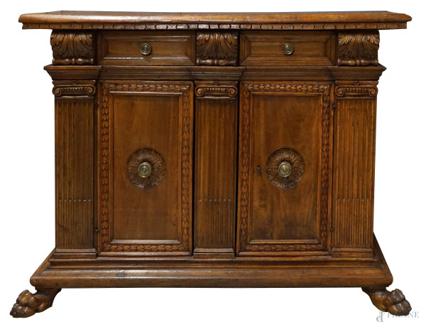 Credenza Rinascimento in noce, piano di linea rettangolare dentellato nella fascia,  particolari scolpiti a motivi di foglie d'acanto, fronte a due cassetti e due sportelli pannellati inquadrati da tre lesene scanalate, due  piedi leonini, finiture in bronzo, cm h 115x164x65, (difetti).