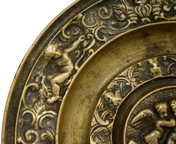 Piatto in bronzo decorato a bassorilievo con tre putti al centro, tesa a grottesche con mascheron...