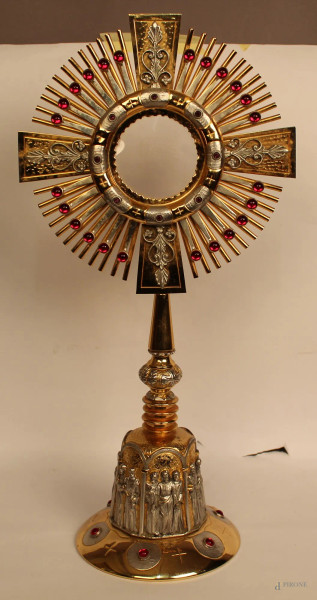 Ostensorio a raggiera, anni 50 in metallo inciso, argentato e dorato, base con figure di santi a rilievo con pietre a color rubino, h. 55 cm. 