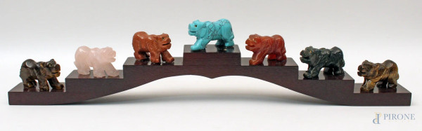 Lotto composto da sette sculture a soggetto di orsi in pietre diverse, poggianti su una base in tek, lunghezza 40 cm.