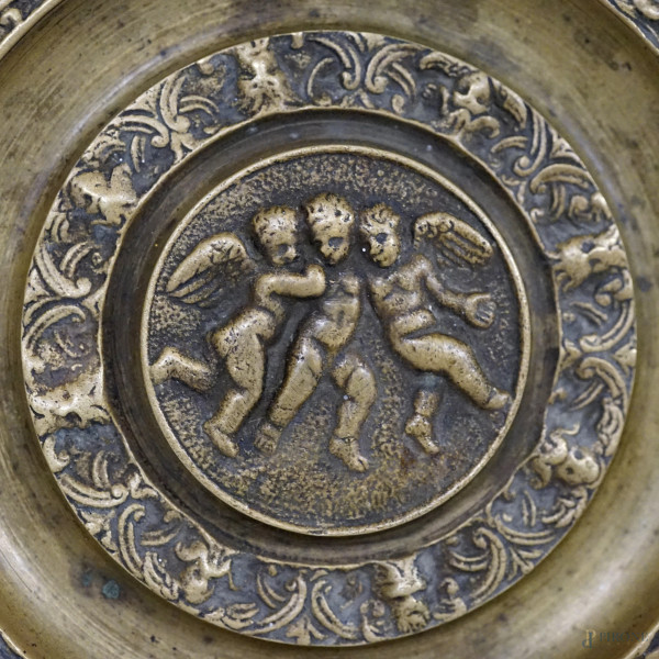 Piatto in bronzo decorato a bassorilievo con tre putti al centro, tesa a grottesche con mascheron...