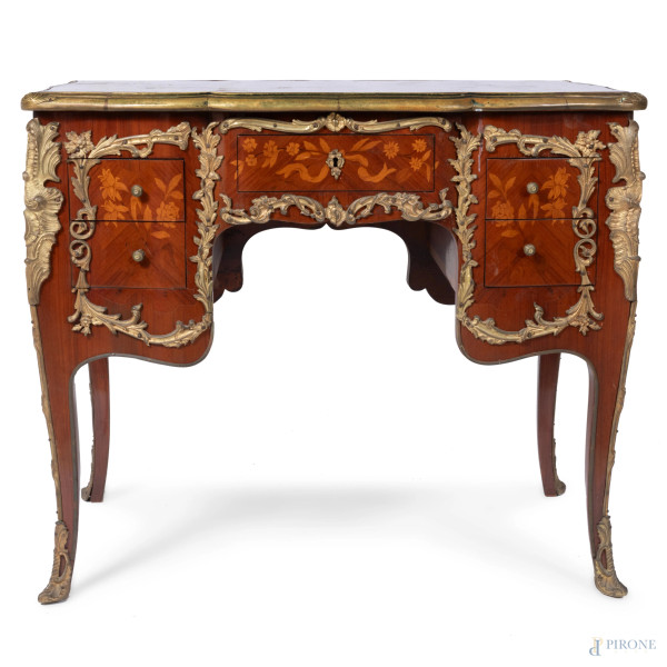 Scrivania in legno impiallacciato, intarsiato in varie essenze e decorato da inserti in bronzo dorato. Gambe a sciabola e fronte scandito da cassetto centrale affiancato da coppia di cassetti in colonna. Piano sagomato con perimetro in bronzo dorato. XX secolo, cm 79x93x54, (graffi, segni di usura)