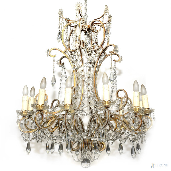 Lampada a sospensione a 12 luci con bracci curvilinei in metallo dorato che sorreggono complesso apparato di pendenti e gocce in cristallo sfaccettato, XX secolo, cm 86x88