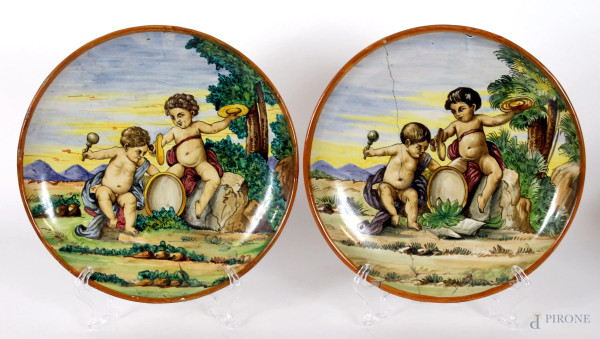 Coppia di piatti in maiolica smaltata, dipinti in policromia con putti musicanti su sfondo di paesaggi, diametro cm. 25, marca sotto la base, (difetti e restauri).