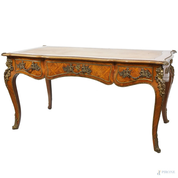 Tavolo scrittoio da centro in legno impiallacciato, metà del XX secolo, piano sagomato con inserto in pelle, tre cassetti nella fascia, quattro gambe mosse, ferramenta in metallo dorato, cm h 79x157x85, (difetti)