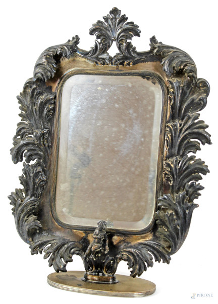 Specchio da tavolo con cornice in argento sbalzato e cesellato a motivi di volute e foglie d'acanto con putto seduto alla base, cm 41x31, inizi XX secolo.