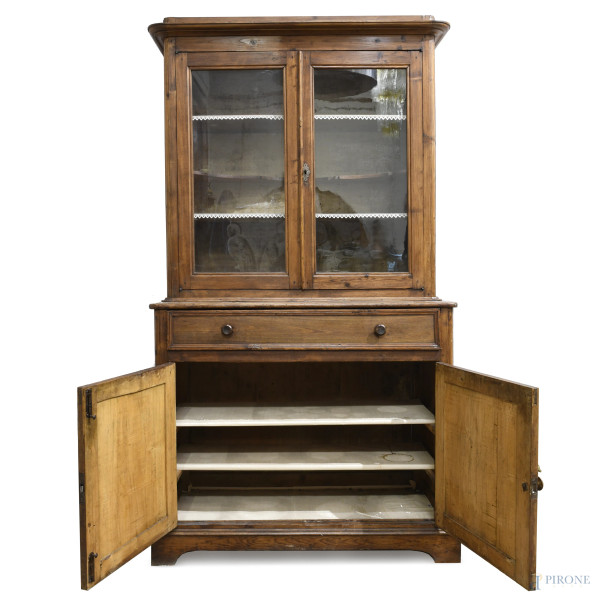Credenza in legno dolce con corpo inferiore scandito da cassettone e doppia anta, corpo superiore...