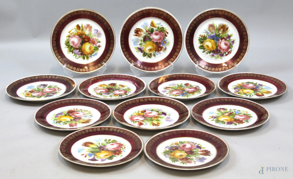 Dodici piatti piani in porcellana policroma a decori floreali, finiture dorate su tesa bordeaux, diametro cm 25, marchio sotto la base
