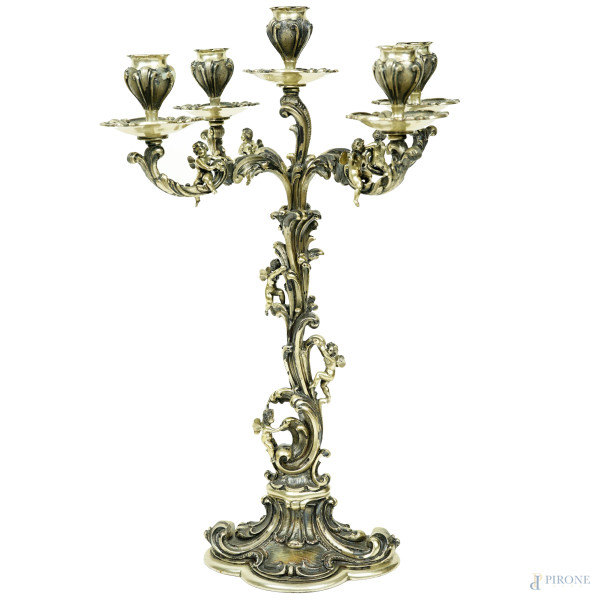 Candelabro in argento a cinque fiamme, bracci riccamente cesellati e sbalzati a volute, foglie e ricci con piccoli putti giocosi, seconda metà XX secolo, cm h 42x29, peso lordo gr. 2320 (appesantito).