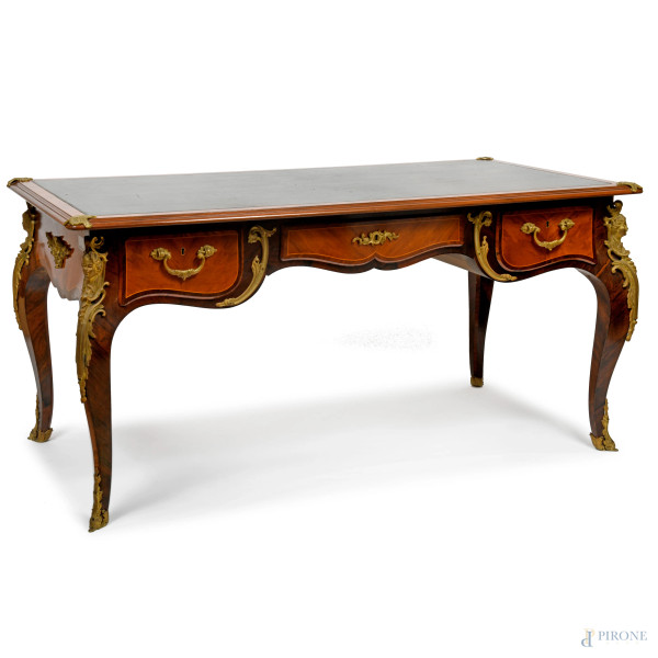Scrivania in stile Luigi XV con ricca decorazione in bronzo dorato e cesellato a motivi rocaille e floreali. Piano con inserto in pelle verde bordato oro. Tre cassetti al fronte. Gambe arcuate terminanti in sabot in bronzo, XX secolo, cm 77x160x79, (difetti)