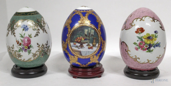 Lotto composto da tre uova in porcellana dipinte a motivi floreali, poggianti su basi in tek, H 18 cm.