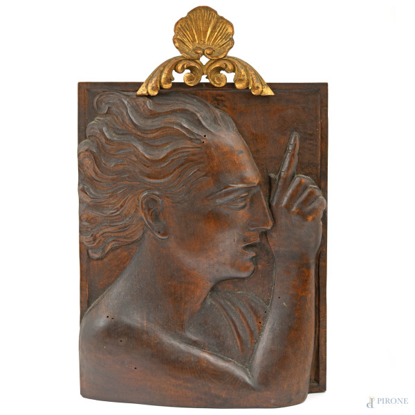 Bassorilevo decorativo in legno intagliato raffigurante un volto maschile di profilo con capelli al vento e il dito puntato verso l'alto. Sormontato da un elemento ornamentale a volute in legno dorato. Inizi del XX secolo, cm 50x32, (difetti).