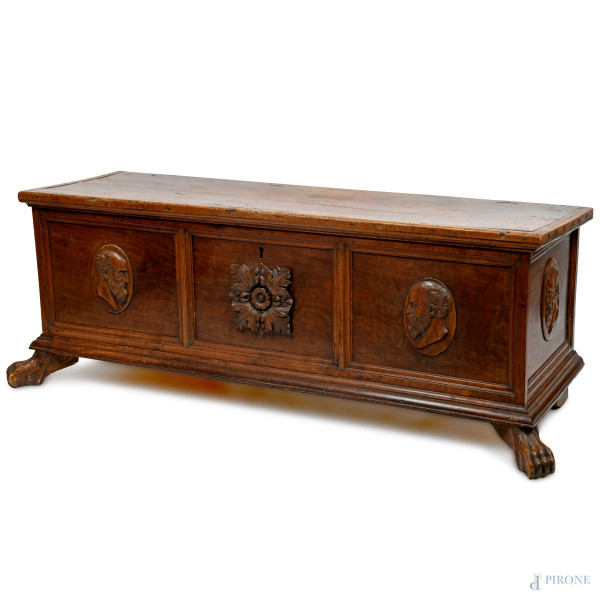 Cassapanca in noce modanato con fronte scandito da tre sezioni contenenti rispettivamente al centro inserto scolpito a foggia di fiore e ai lati medaglioni in legno scolpito con teste virili, presenti anche ai fianchi.  XVI-XVII secolo, cm 59x159x65, (difetti)