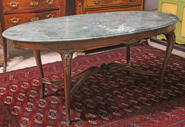 Tavolo di forma ovale in noce, piano in marmo verde, gambe mosse riunite da traversa, finiture in bronzo, cm h80x189x94,5