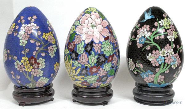 Lotto composto da tre uova in ottone e smalto cloisonn&#232;, a decori floreali, poggianti su basi in tek, H 26 cm.