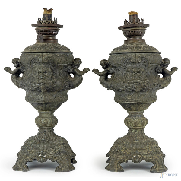 Coppia di lampade da tavolo in metallo sbalzato e cesellato, decori a mascheroni, prese a guisa di putti, cm h 40, fine XIX secolo.